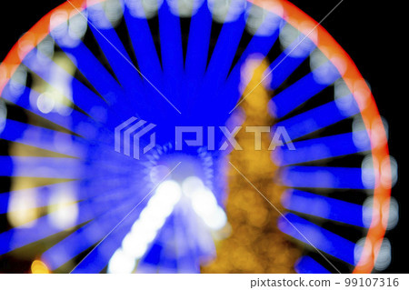 Ferris wheel decorated with blue illumination and a large Christmas tree decorated with yellow illumination on a black background at night. Beautiful New Year and Christmas holiday blurred background Ferris wheel decorated with blue illumination and a large Christmas tree decorated with yellow illumination on a black background at night. Beautiful New Year and Christmas holiday blurred background 99107316