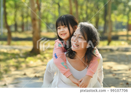 Smiling middle age grandmother giving piggyback ride to cute little granddaughter during walking on in the park 99107706