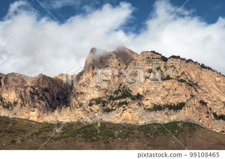 Beautiful long pointed rock is illuminated by the sun. Sunny mountain landscape with high sharp rockies under white clouds. Panoramic view of sharp rocks and peace top in blue cloudy sky. 99108465