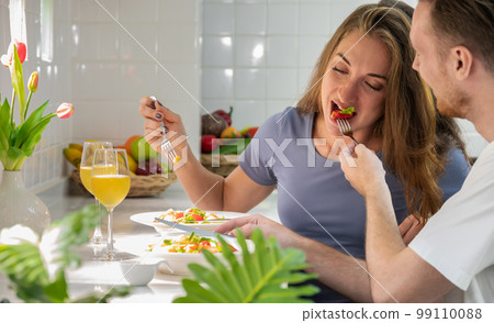 Happy couple eating salad in kitchen at home. Young man giving strawberry to his girlfriend. . Happy couple eating salad in kitchen at home. Young man giving strawberry to his girlfriend. . 99110088