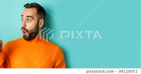 Close-up of young bearded man showing phone screen and looking amazed, wearing orange sweater, standing against studio background Close-up of young bearded man showing phone screen and looking amazed, wearing orange sweater, standing against studio background 99120051