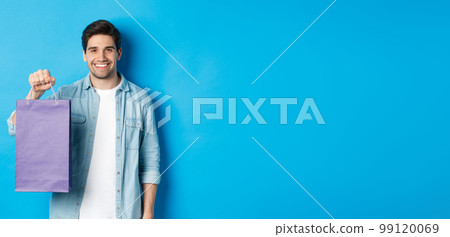 Concept of shopping, holidays and lifestyle. Young handsome man holding paper bag with present and smiling, standing over blue background Concept of shopping, holidays and lifestyle. Young handsome man holding paper bag with present and smiling, standing over blue background 99120069