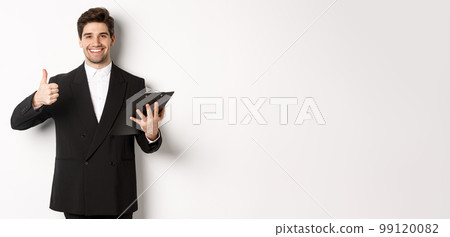 Portrait of confident businessman in black suit, holding clipboard with documents and showing thumb-up in approval, praise good job, standing against white background 99120082
