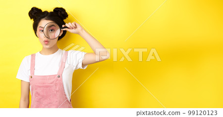 Silly glamour lady with bright makeup, look through magnifying glass and pouting, she is watching you, standing on yellow background 99120123