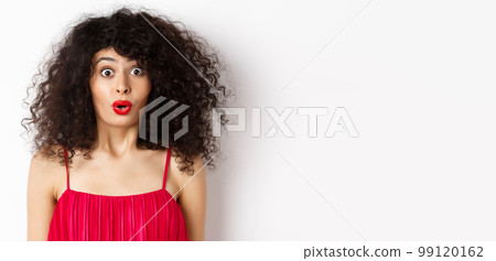 Close-up of surprised caucasian woman with red lips and curly hairstyle, saying wow and looking amazed, standing in red dress over white background Close-up of surprised caucasian woman with red lips and curly hairstyle, saying wow and looking amazed, standing in red dress over white background 99120162