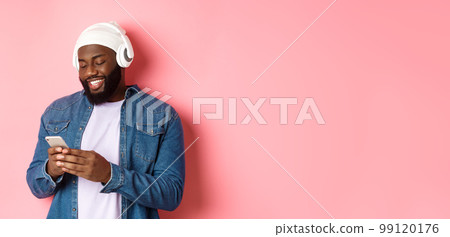 Handsome stylish Black man listening music in headphones and writing message on smartphone, smiling happy, standing over pink background Handsome stylish Black man listening music in headphones and writing message on smartphone, smiling happy, standing over pink background 99120176