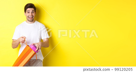Concept of holidays and celebration. Happy and surprised man take out gift and looking at camera, standing over yellow background 99120205