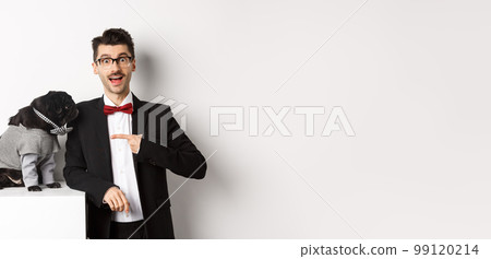 Cheerful guy standing with his cute pug, pointing finger at dog wearing party costume, posing over white background 99120214
