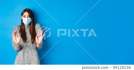 Covid-19, pandemic lifestyle concept. No thank you. Reluctant and worried girl in medical mask asking non-masker to stay away, showing block gesture, tell stop, blue background Covid-19, pandemic lifestyle concept. No thank you. Reluctant and worried girl in medical mask asking non-masker to stay away, showing block gesture, tell stop, blue background 99120236