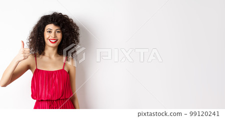 Cheerful woman with red lips and elegant dress, showing thumb up and smiling, recommending product, standing over white background Cheerful woman with red lips and elegant dress, showing thumb up and smiling, recommending product, standing over white background 99120241