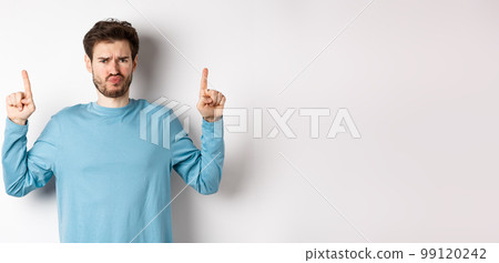 Disappointed male model frowning and sulking, pointing fingers up with negative emotion, dislike promo offer, standing over white background Disappointed male model frowning and sulking, pointing fingers up with negative emotion, dislike promo offer, standing over white background 99120242