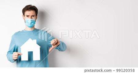 Covid and real estate concept. Disappointed young man in medical mask showing paper house cutout and thumb down, dislike broker agency, standing displeased over white background 99120358