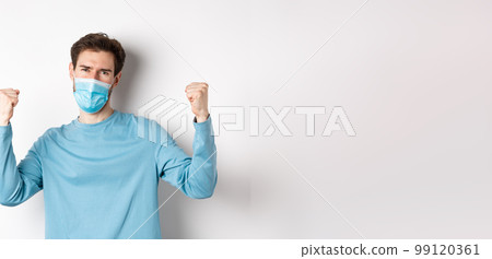 Covid-19, pandemic and social distancing concept. Happy young man in medical mask winning, screaming yes with satisfaction and raising hands up, celebrating victory, white background 99120361