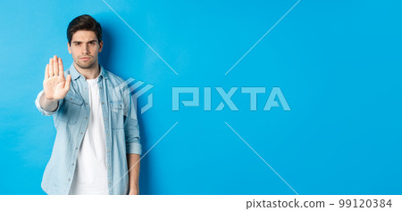 Serious man frowning and saying no, extending hand to shop stop sign, prohibit action, standing against blue background Serious man frowning and saying no, extending hand to shop stop sign, prohibit action, standing against blue background 99120384