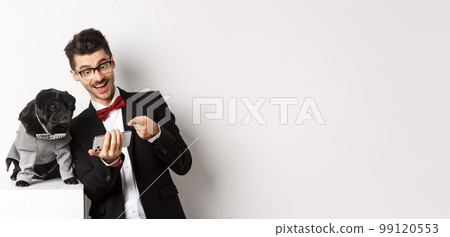 Happy dog owner showing something to pet on mobile phone, man and pug wearing fancy costumes, standing over white background Happy dog owner showing something to pet on mobile phone, man and pug wearing fancy costumes, standing over white background 99120553