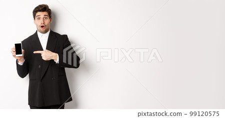 Portrait of attractive businessman in black suit, looking surprised and pointing finger at smartphone sreen, standing over white background Portrait of attractive businessman in black suit, looking surprised and pointing finger at smartphone sreen, standing over white background 99120575