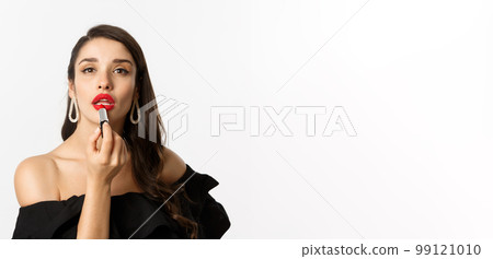 Close-up of beautiful woman applying red lipstick on lips, looking at camera like mirror, standing in black dress over white background 99121010