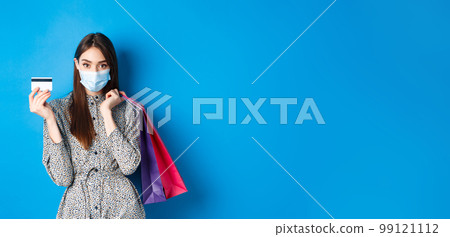 Covid-19, pandemic and lifestyle concept. Stylish woman wearing medical mask on shopping as preventive measure from coronavirus, showing plastic credit card and holding bags, blue background 99121112