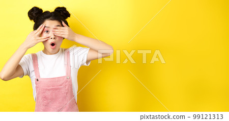 Excited asian teen girl covering eyes with hands and peeking through fingers, gasping amazed at camera, standing in summer clothes on yellow background Excited asian teen girl covering eyes with hands and peeking through fingers, gasping amazed at camera, standing in summer clothes on yellow background 99121313