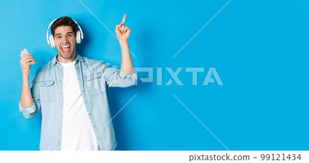 Joyful handsome man dancing with smartphone, listening music in headphones and pointing finger up, standing over blue background Joyful handsome man dancing with smartphone, listening music in headphones and pointing finger up, standing over blue background 99121434