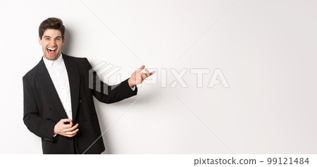 Portrait of happy man dancing at party, playing on invisible guitar and laughing, standing in black suit against white background 99121484