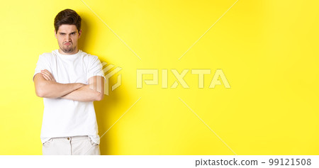 Offended and mad guy, frowning and sulking, holding hands crossed on chest, standing angry against yellow background 99121508
