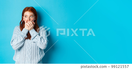 Shocked and anxious redhead girl staring left scared, covering mouth and gasping, standing over blue background in sweater Shocked and anxious redhead girl staring left scared, covering mouth and gasping, standing over blue background in sweater 99121557