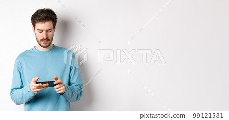 Male model playing video game on smartphone, looking serious at mobile screen, standing in sweatshirt over white background 99121581
