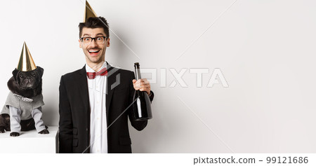 Happy young man celebrating with dog. Cute black pug and hipster wearing birthday party cones, guy holding champagne, standing over white background Happy young man celebrating with dog. Cute black pug and hipster wearing birthday party cones, guy holding champagne, standing over white background 99121686