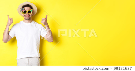Concept of tourism and lifestyle. Image of smiling tourist showing thumbs-up, enjoying trip and recommending, wearing summer hat and sunglasses, yellow background Concept of tourism and lifestyle. Image of smiling tourist showing thumbs-up, enjoying trip and recommending, wearing summer hat and sunglasses, yellow background 99121687