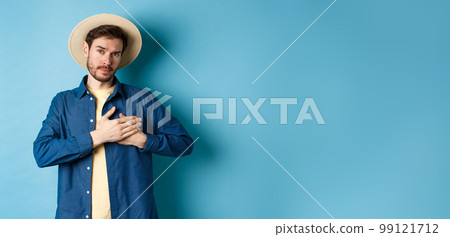 Heartfelt young woman in tourist hat, holding hands on heart and looking thankful and serious, standing on blue background Heartfelt young woman in tourist hat, holding hands on heart and looking thankful and serious, standing on blue background 99121712