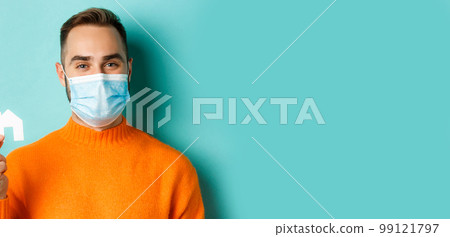 Real estate and coronavirus pandemic concept. Close-up of adult man in medical mask holding small paper house maket and smiling, searching for apartment, light blue background 99121797