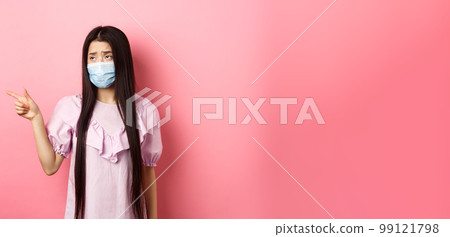 Coronavirus, quarantine and lifestyle concept. Sad and gloomy asian woman in medical mask pointing, looking left upset, showing unfair thing, standing on pink background 99121798