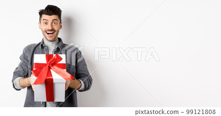 Surprised young man smiling excited, holding big gift box on valentines day holiday, receive surprise present, standing amazed on white background Surprised young man smiling excited, holding big gift box on valentines day holiday, receive surprise present, standing amazed on white background 99121808