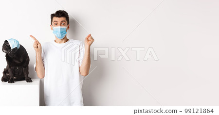 Covid-19, animals and quarantine concept. Young man and black dog wearing medical masks, pug looking at upper left corner and owner showing promo offer, rejoicing 99121864