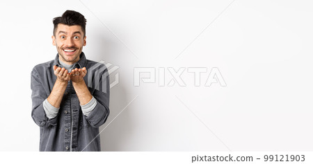 Lovely young man sending air kiss at camera and smiling. Hopeless romantic showing his love, holding something in hands, standing on white background 99121903