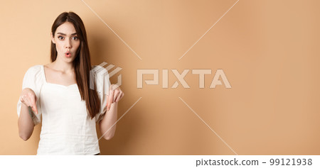 Wow amazing. Impressed young woman look astonished by product, pointing fingers down at logo, standing on beige background 99121938