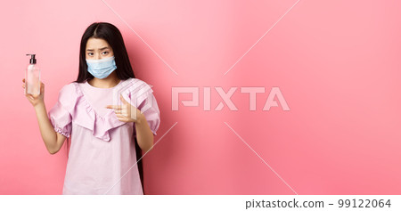 Healthy people and covid-19 pandemic concept. Skeptical asian woman frowning, wearing face mask, pointing at bottle of hand sanitizer, standing against pink background Healthy people and covid-19 pandemic concept. Skeptical asian woman frowning, wearing face mask, pointing at bottle of hand sanitizer, standing against pink background 99122064