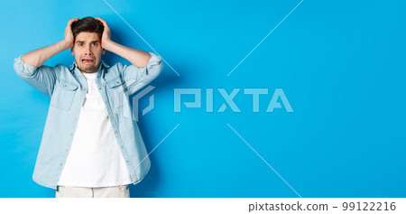 Image of man in panic holding hands on head, looking frustrated and anxious, standing against blue background Image of man in panic holding hands on head, looking frustrated and anxious, standing against blue background 99122216