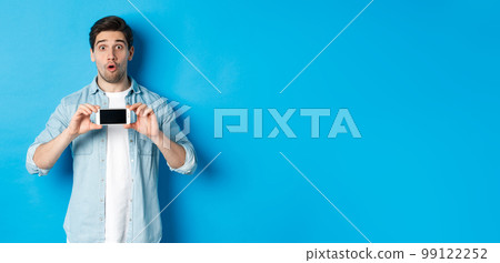 Surprised guy showing mobile phone screen and looking impressed, standing over blue background 99122252