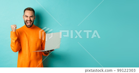 Freelance and technology concept. Lucky man winner celebrating, winning online, showing fist pump and holding laptop, standing over light blue background Freelance and technology concept. Lucky man winner celebrating, winning online, showing fist pump and holding laptop, standing over light blue background 99122303