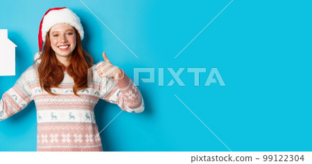 Holiday promos and real estate concept. Satisfied female model with red wavy hair, wearing santa hat and sweater, showing paper house model and thumbs-up, blue background Holiday promos and real estate concept. Satisfied female model with red wavy hair, wearing santa hat and sweater, showing paper house model and thumbs-up, blue background 99122304