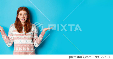 Image of excited redhead girl react to good news, looking surprised, spread hands sideways and smiling, standing in winter sweater against blue background Image of excited redhead girl react to good news, looking surprised, spread hands sideways and smiling, standing in winter sweater against blue background 99122305