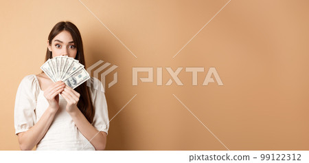 Excited girl showing dollar bills and raising eyebrows amazed, making money, standing on beige background 99122312
