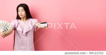 Excited teen asian girl stretch out hands with dollar bills and plastic credit card, look at money with amazement, standing on pink background 99122332