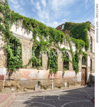 Old abandoned building, with stone wall, and wrought iron windows, covered with climber plants, in cobblestone alley 99122362