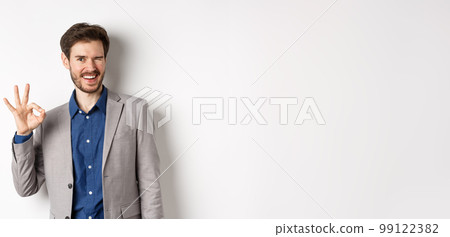 Everything okay. Cheerful businessman in suit winking and showing OK sign, smiling assertive, guarantee quality, praise good choice, standing on white background Everything okay. Cheerful businessman in suit winking and showing OK sign, smiling assertive, guarantee quality, praise good choice, standing on white background 99122382