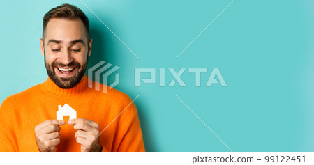 Real estate concept. Happy young man searching for home rent, holding house paper maket and smiling, standing over blue background 99122451