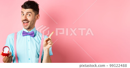 Valentines day. Funny guy with moustache and bow-tie, showing engagement ring and pointing finger at upper right corner, standing on pink background Valentines day. Funny guy with moustache and bow-tie, showing engagement ring and pointing finger at upper right corner, standing on pink background 99122488