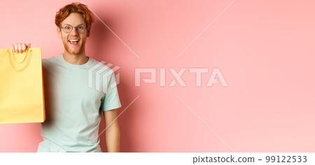 Cheerful redhead man in t-shirt, showing shopping bag and smiling, standing over pink background 99122533